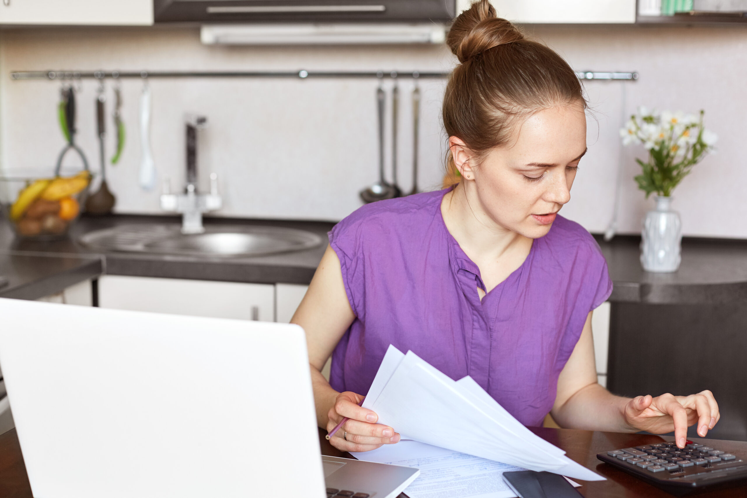 Busy working female freelancer makes business report, calculates figures and holds documents, works on laptop computer, connected to wireless internet at home, sits against kitchen interior.